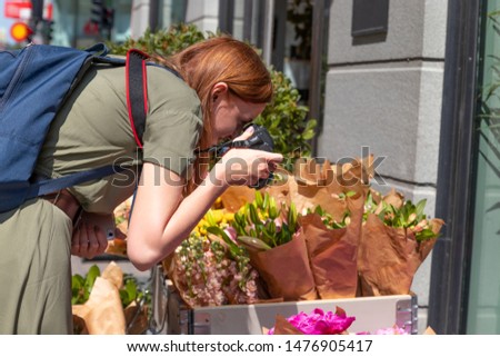 Young blonde girl photographer makes a photo of assorted flowers bouquets in a shop on the street