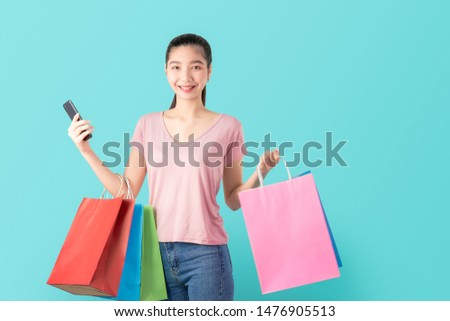 Smiling asian woman casual style holding smartphone and shopping bags on light blue background.