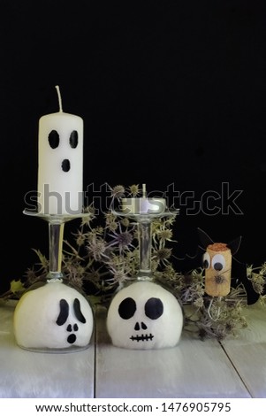 Charming stylish ghosts from glass goblets and white wax candle in dry thistle and bat from cork and black cardboard on natural wooden surface against black wall.