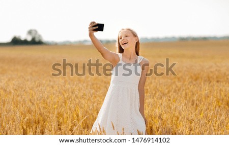 technology, summer and people concept - smiling young girl in white dress taking selfie by smartphone on cereal field