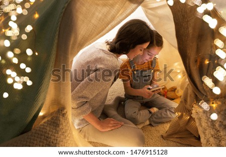 family, hygge and technology concept - happy mother and little daughter with smartphone in kids tent at night at home
