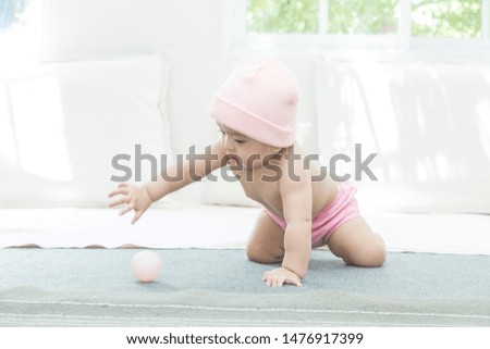 Happy asian baby wearing pink wool hat playing pink ball on grey mattress in living room with natural lighting