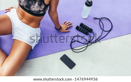 Cropped shot of a woman in fitness wear relaxing after workout sitting on a yoga mat with mobile phone, water bottle and skipping rope beside her. Royalty-Free Stock Photo #1476944936