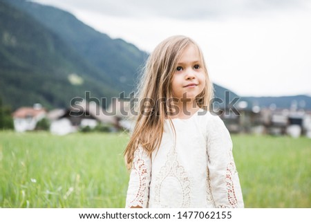 A little blond girl in a white dress is playing on a nature near the mountains on the grass.