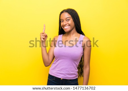 African American teenager girl with long braided hair over isolated yellow wall showing and lifting a finger in sign of the best