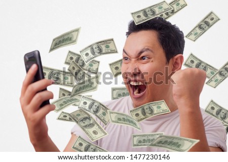 Portrait of happy successful young Asian billionaire man smiling happily under rain of money from his phone. Wealth investment internet online business concept