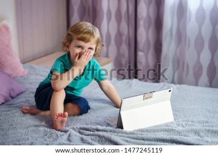 Golden-haired, charming child watches cartoons on a tablet computer while sitting on a bed in the parent’s bedroom. Interesting pastime alone.
