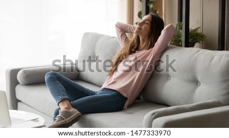 Serene young woman pretty relaxing on couch in living room. Calm female freelancer crossing hands behind head, closing eyes and taking a break during remote work day at home, stress free concept. Royalty-Free Stock Photo #1477333970