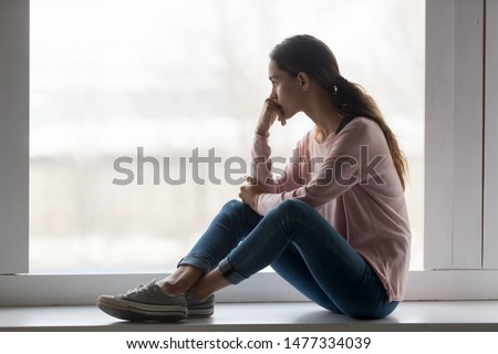 Young mixed race thoughtful woman sitting on window sill at home, holding head in hand, looking outside blurred window, remembering sad moments, feeling lonely, thinking over problem or mistakes. Royalty-Free Stock Photo #1477334039