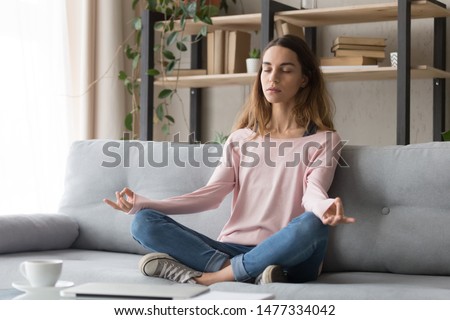 Carefree calm millennial freelance girl sitting on couch in lotus position and mudra gesture, relaxing, doing yoga practice. Young serene woman meditating, visualizing, healthy lifestyle concept. Royalty-Free Stock Photo #1477334042