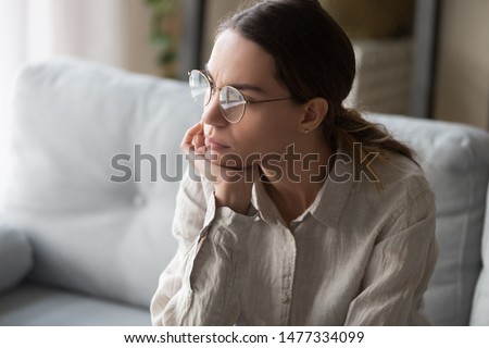 Upset young mixed race woman in eyeglasses sitting on couch in living room, holding head in hand, looking outside, thinking over problems, making difficult decision, feeling tired, exhausted. Royalty-Free Stock Photo #1477334099