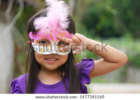 The portrait picture of a lovely little Asian girl wearing a mask, making a victory sign hands and smiling in the garden background.