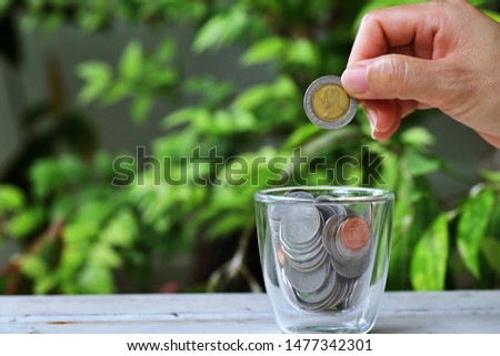 The woman's hand putting money coins to glass jar with green leaf background.