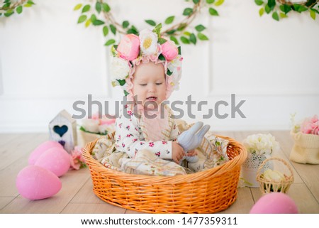 Cute little girl in a dress with a flower print and a hat with flowers in the Easter decorations in the studio. Little girl with easter eggs and flowers in a spring studio
