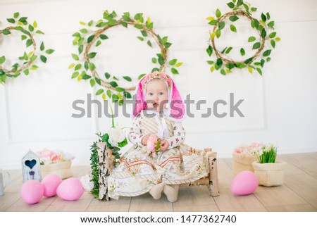 Cute little girl in a dress with a flower print and a bunny hat in the Easter decorations in the studio. Little girl with easter eggs and flowers in a spring studio
