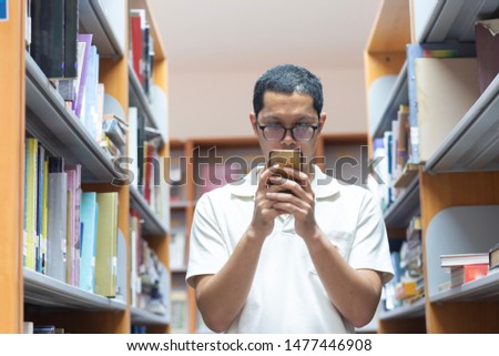 The man is standing between the book shelfs and looking to the smartphone, technology, search engine, library, text book, learning, university, academic concept.