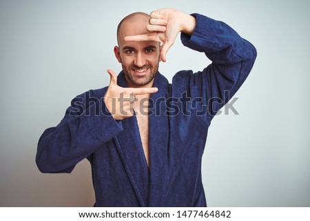 Young man wearing blue bathrobe, relaxed lifestyle over isolated background smiling making frame with hands and fingers with happy face. Creativity and photography concept.