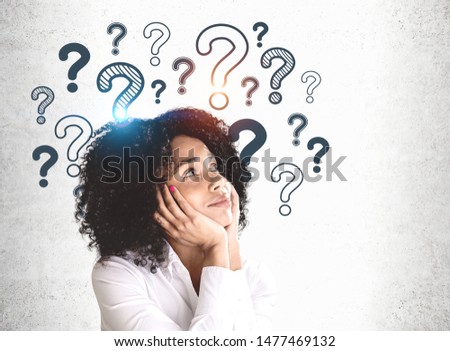Smiling young African American woman in white shirt looking at question marks drawn on concrete wall. Concept of curiosity and education Royalty-Free Stock Photo #1477469132