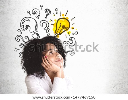 Smiling young African American woman in white shirt looking at question marks and lightbulb drawn on concrete wall. Concept of good idea. Mock up Royalty-Free Stock Photo #1477469150
