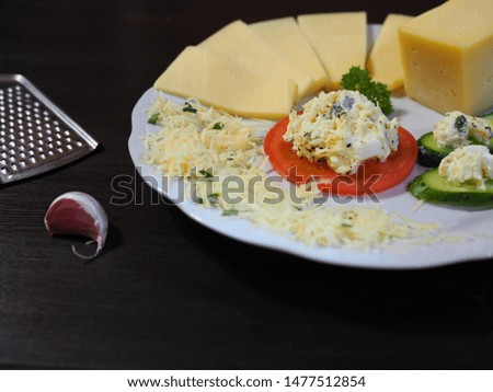 Slices and a piece of fresh different cheese, grated cheese, tomato and cucumber slices with cheese filling, parsley branch in a white plate on the background of grater and garlic on the table.