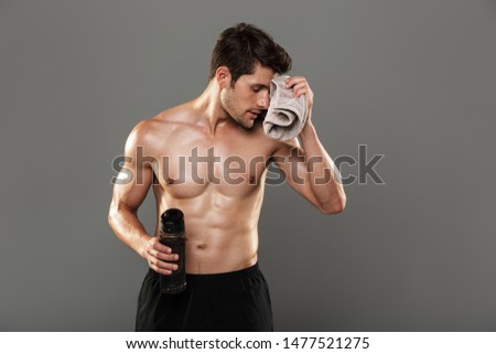 Photo of a tired handsome young strong sportsman isolated over grey wall background holding towel and bottle with water.