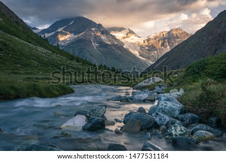 Sunrise in Swiss Alps, near Tasch. A river runs through meadows and pastures. Distant mountains and glaciers, as well as clouds in the sky, catch the orange light of the morning.