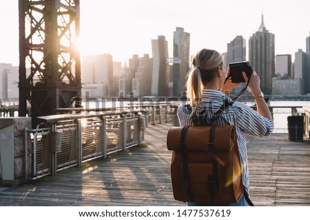 Back view of stylish female tourist with traveling backpack standing on American urban setting and clicking pictures of Manhattan landmark using retro instant camera, concept of photography hobby Royalty-Free Stock Photo #1477537619