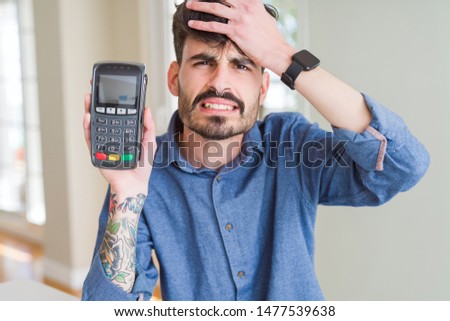 Young man holding dataphone point of sale as payment stressed with hand on head, shocked with shame and surprise face, angry and frustrated. Fear and upset for mistake.