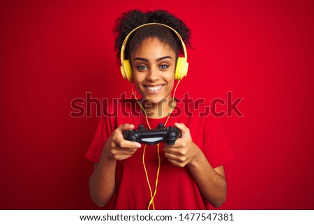 Afro woman playing video game using joystick and headphones over isolated red background with a happy face standing and smiling with a confident smile showing teeth