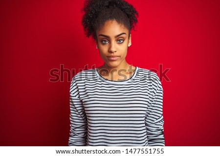African american woman wearing navy striped t-shirt standing over isolated red background with serious expression on face. Simple and natural looking at the camera.