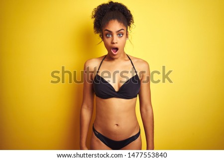 African american woman on vacation wearing bikini standing over isolated yellow background In shock face, looking skeptical and sarcastic, surprised with open mouth