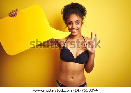 African american woman wearing bikini holding speech bubble over isolated yellow background doing ok sign with fingers, excellent symbol