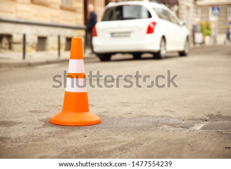 Repair work sign restricting traffic. White-orange plastic striped cone warning of danger. Tightening in traffic. Street of the old city with cars. Roadway repair. Poor asphalt on the road. Attention 