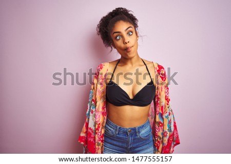 American woman on holiday wearing bikini and colorful dress over isolated pink background making fish face with lips, crazy and comical gesture. Funny expression.