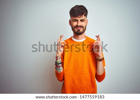 Young man with tattoo wearing orange sweater standing over isolated white background gesturing finger crossed smiling with hope and eyes closed. Luck and superstitious concept.