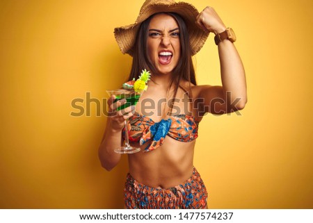 Woman on vacation wearing bikini and hat drinking cocktail over isolated yellow background annoyed and frustrated shouting with anger, crazy and yelling with raised hand, anger concept