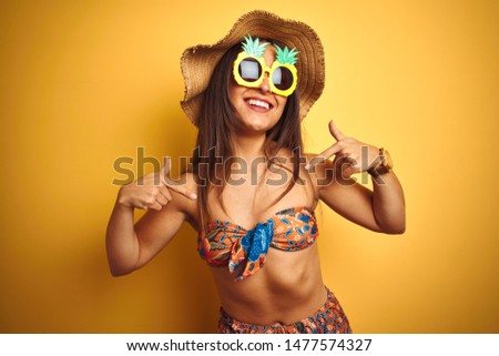 Woman on vacation wearing bikini and pineapple sunglasses over isolated yellow background looking confident with smile on face, pointing oneself with fingers proud and happy.