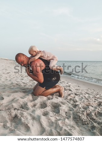 dad and daughter are playing on the beach, he took her on his shoulders. Baby in a cute pink jumpsuit and hat