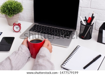 Colored atmosphere. Woman holding red cup with coffee in front of laptop with blank screen on office table