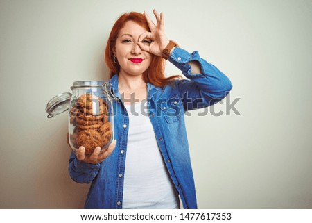 Young beautiful redhead woman holding jar of cookies over white isolated background with happy face smiling doing ok sign with hand on eye looking through fingers