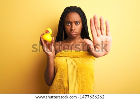 African american woman wearing shower towel holding toy duck over isolated yellow background with open hand doing stop sign with serious and confident expression, defense gesture