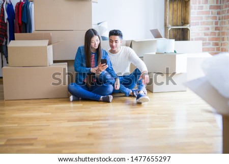 Young asian couple sitting on the floor of new apartment arround cardboard boxes, using smartphone and smiling at new home