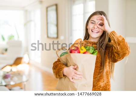 Beautiful young girl holding paper bag of fresh groceries with happy face smiling doing ok sign with hand on eye looking through fingers