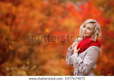 happy girl with blonde curls and a beautiful smile in a lilac coat, red hat and red scarf holds a bouquet of autumn leaves in a beautiful red-gold autumn forest