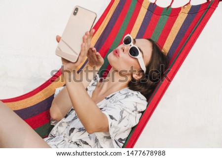 Image of a young beautiful woman at the beach posing on a hammock take a selfie by mobile phone blowing kisses.