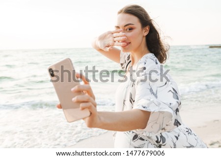 Photo of a pleased happy young beautiful woman at the beach walking in a beautiful sunny morning take a selfie by mobile phone covering face.