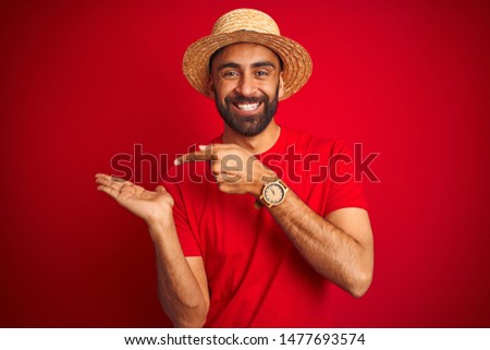 Young handsome indian man wearing t-shirt and hat over isolated red background amazed and smiling to the camera while presenting with hand and pointing with finger.