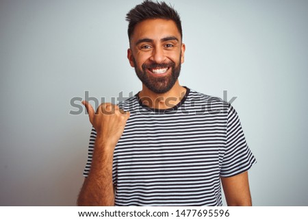 Young indian man wearing black striped t-shirt standing over isolated white background smiling with happy face looking and pointing to the side with thumb up.