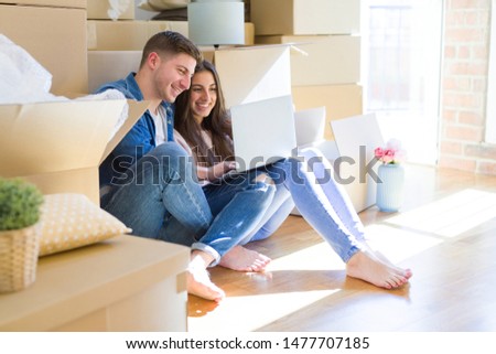 Young couple sitting on the floor of new house around cardboard boxes using laptop, smiling happy for new apartment