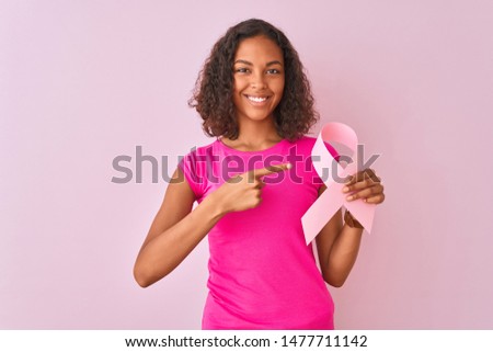 Young brazilian woman holding cancer ribbon standing over isolated pink background very happy pointing with hand and finger
